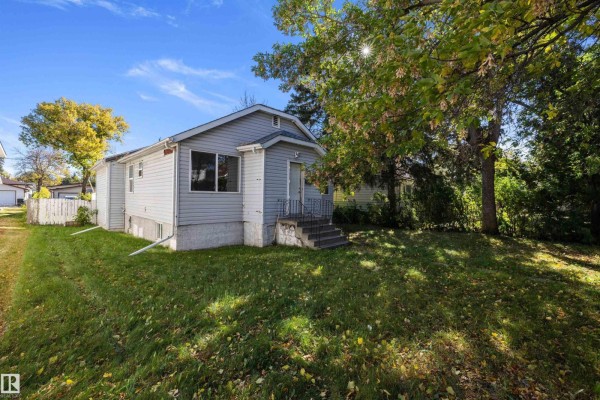 Angle view of the bungalow's left side with lawn, trees, and a concrete foundation. - 12043 87 St Nw, Edmonton