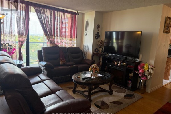 Living room with sofas, coffee table, TV, fireplace, and view through red patterned curtains. - 1205 - 17 Knightsbridge Road, Brampton