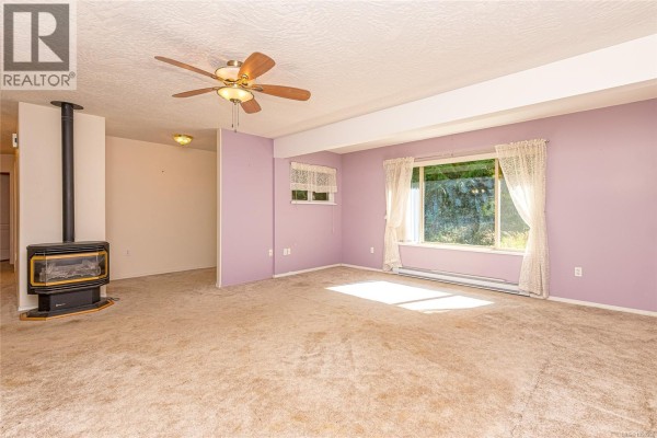 Living room with beige carpet, lavender walls, a wood-burning stove, and a large window. - 1205 Corcan Rd, Qualicum Beach