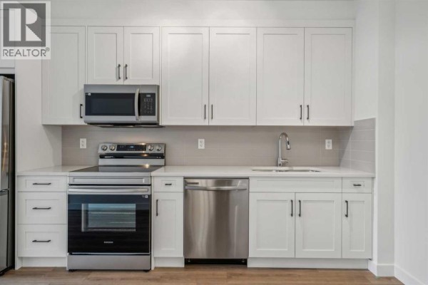 Kitchenette with white cabinetry, stainless steel appliances including a dishwasher, and light grey backsplash tiles. - 1206, 14910 1 Street Nw, Calgary