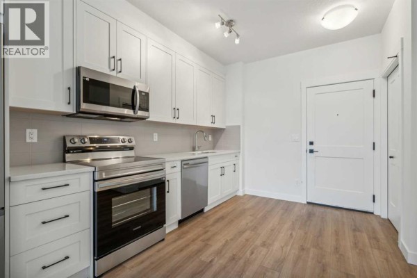 Compact kitchen with white cabinets, stainless steel appliances, and a view of the entryway and adjacent living space. - 1206, 14910 1 Street Nw, Calgary