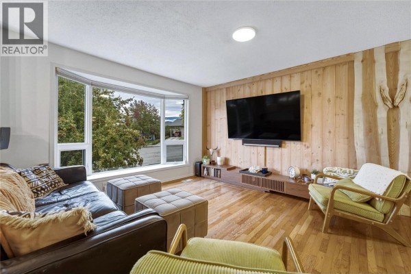 Living room with a large bay window, dark leather sofa, and wood-paneled wall. - 1206 39b Avenue, Vernon