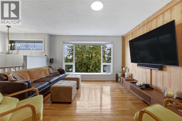 Living room featuring a brown leather sofa, bay window with green trees view, and wood paneling. - 1206 39b Avenue, Vernon