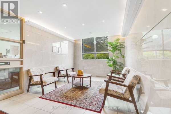 Seating area in lobby with chairs, rug, coffee table, and window overlooking greenery. - 1207 - 10 Stonehill Court, Toronto
