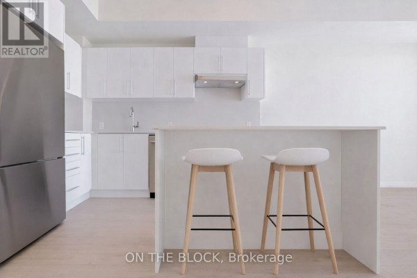 Modern white kitchen with island, two bar stools, and stainless steel refrigerator. - 1207 - 27 Korda Gate, Vaughan