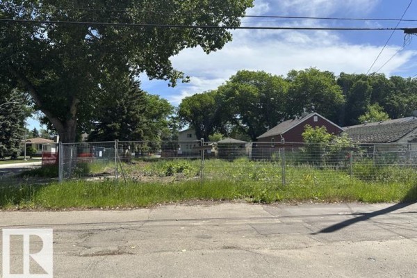Fenced lot with a sign, houses, and trees under a clear blue sky. - 12072 95 St Nw, Edmonton