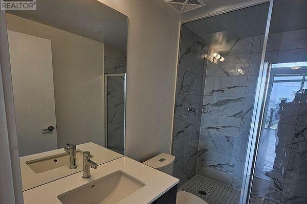 A modern bathroom with a white vanity, marble-patterned tile shower, and toilet. - 1209 - 1350 Ellesmere Road, Toronto