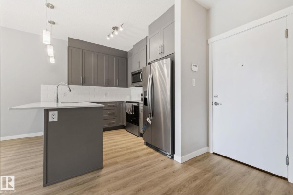 A spacious apartment living area and kitchen with grey cabinets, a white island, and a window view. - #121 812 Welsh Dr Sw, Edmonton