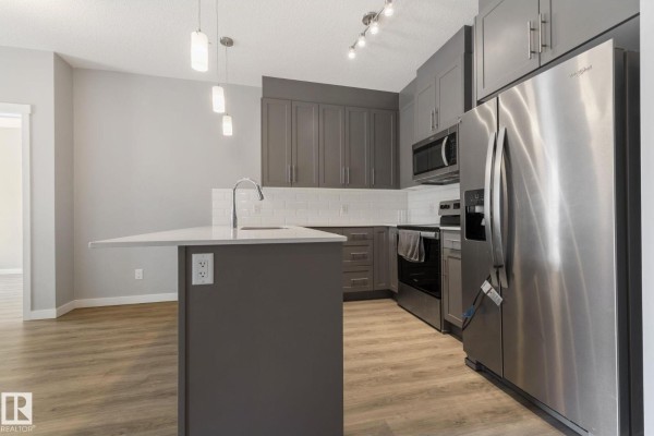 This view showcases the kitchen's dark grey cabinets, a stainless steel dishwasher, and the light wood laminate flooring. - #121 812 Welsh Dr Sw, Edmonton