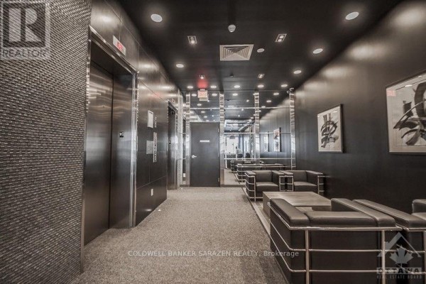 Modern building lobby with dark walls, elevators, mirrored surfaces, grey carpet, and black leather seating. - 1210 - 224 Lyon Street, Ottawa