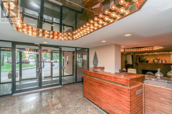 Interior view of the apartment building lobby with glass doors, a wooden reception desk, and decorative lighting. - 1213 - 2871 Richmond Road W, Ottawa