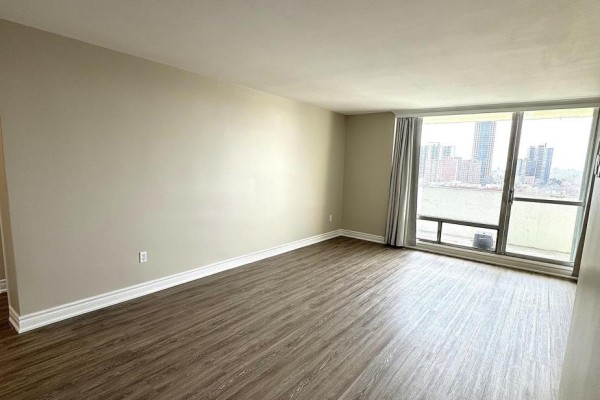 Empty living room with beige walls, dark wood laminate flooring, and city view through large windows. - 1214 - 1 Massey Square, Toronto