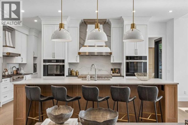 Kitchen island with wooden base and white countertop, featuring bar stools and pendant lights, with appliances behind. - 1217 38 Avenue Sw, Calgary
