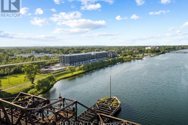 Aerial view from a high vantage point showing apartment buildings on a riverbank with greenery and trees. - 122 - 350 Prince Charles Drive S, Welland
