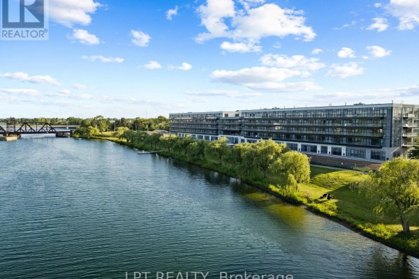 Wide aerial view of apartment buildings situated along a river, with a treed landscape and distant horizon. - 122 - 350 Prince Charles Drive S, Welland