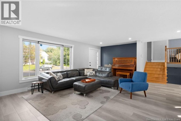 Living room with a dark grey sectional sofa, blue armchair, piano, and staircase. - 122 Dolbeau Street, Dieppe