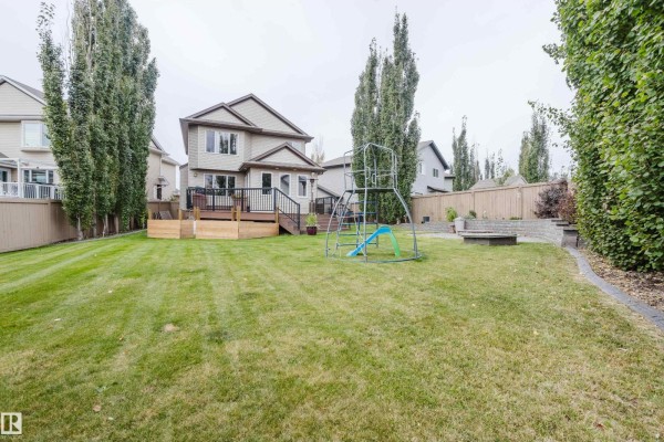 Rear view of a spacious green backyard with a playground, wooden deck, and fence. - 12207 171 Av Nw, Edmonton