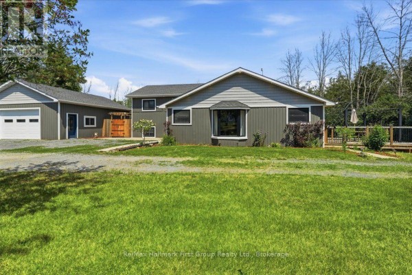 Front exterior view of a grey house with a bay window, wide green lawn, and wooden deck. - 1233 Lazier Road, Tyendinaga