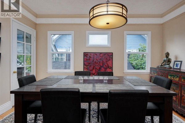 Dining room with dark wood table, black chairs, decorative cabinet, and windows overlooking the exterior. - 1240 Victoria Ave, Oak Bay