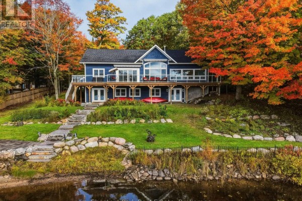 Exterior view of a blue house with a deck, surrounded by vibrant autumn trees and water. - 1252 Winders Bay Road, Lake Of Bays