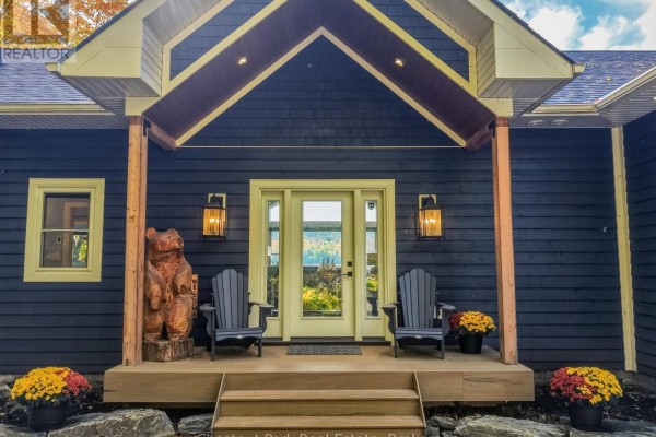 Front porch entrance of a dark blue house with wood accents, a bear carving, and two chairs. - 1252 Winders Bay Road, Lake Of Bays