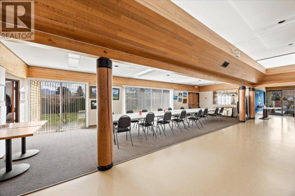 Large community room with long tables, chairs, wood beam ceiling, and a park view. - 1255 Raymer Avenue Unit# 493, Kelowna