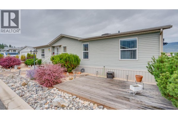 Side exterior of a light grey mobile home showing a wooden deck and decorative landscaping. - 12560 Westside Road Unit# 15, Vernon