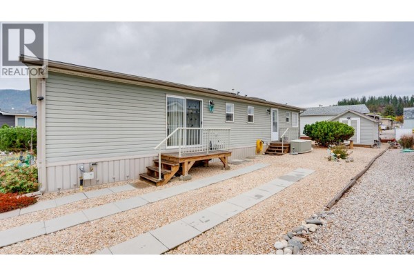 Rear exterior of a light grey mobile home with two wooden staircases and gravel pathways. - 12560 Westside Road Unit# 15, Vernon