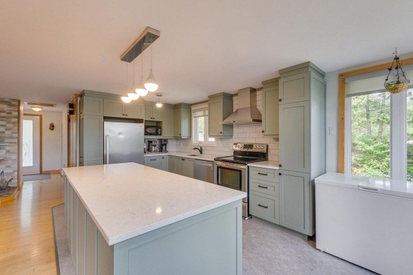 A spacious kitchen with sage green cabinets, a white quartz island, stainless steel appliances, and a view of the outdoors. - 126-129 Ch. Richard, Bouchette