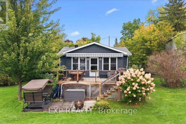 Side view of a dark blue clapboard house with a wooden deck and stairs, surrounded by green lawn and trees. - 126 Gardiner Shore Road, Beckwith