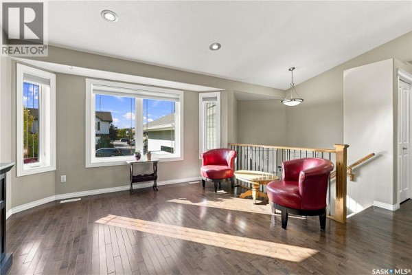 Living room with hardwood floors, taupe walls, two red chairs, and a staircase. - 126 Lamarsh Road, Saskatoon