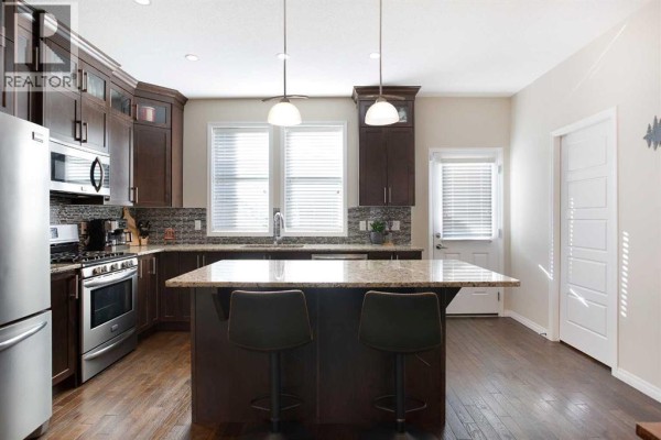Kitchen with dark wood cabinets, granite countertops, stainless steel appliances, and an island with bar stools. - 126 Williamstown Park Nw, Airdrie