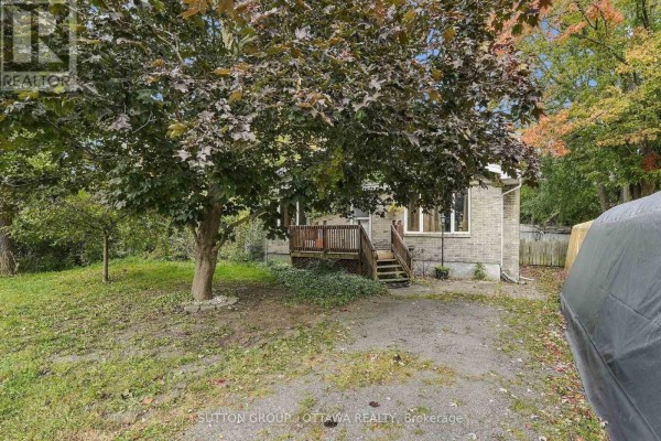 Driveway view of the house entrance, framed by a large deciduous tree and surrounding foliage. - 12615 County 28 Road, South Dundas