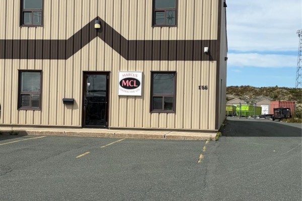 Close-up view of a tan and brown metal commercial building entrance with a glass door and office signs. Parking lot in foreground. - 1266 Kenmount Road Unit#c, Paradise