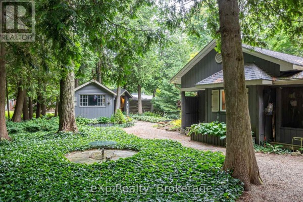 A grey cottage with a covered porch and wooden deck, surrounded by trees and gravel. - 127 Paisley Drive, Chatsworth