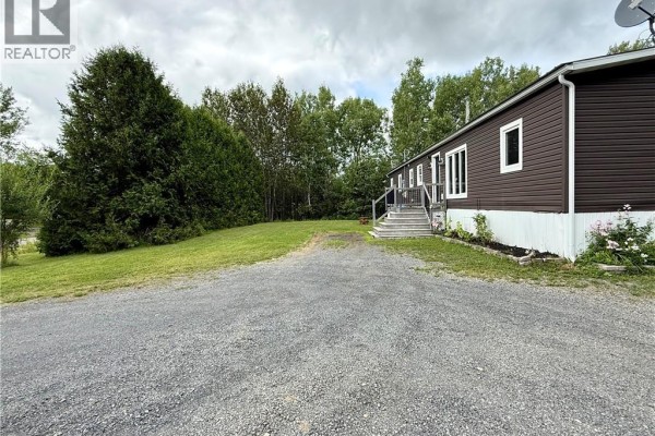 A wider view of the property, showing the mobile home's side, lawn, gravel driveway, and surrounding trees. - 1270 Back Lake Road, New Denmark