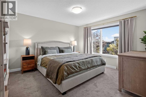 Primary bedroom with a king-size bed, grey carpet, large window with curtains, and wooden dresser. - 1270 Walden Drive Se, Calgary