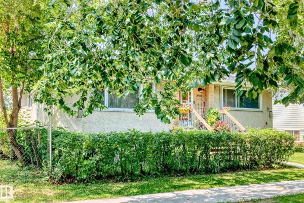 A partial view of the duplex exterior obscured by lush green tree branches and hedges. - 12718 126 St Nw, Edmonton