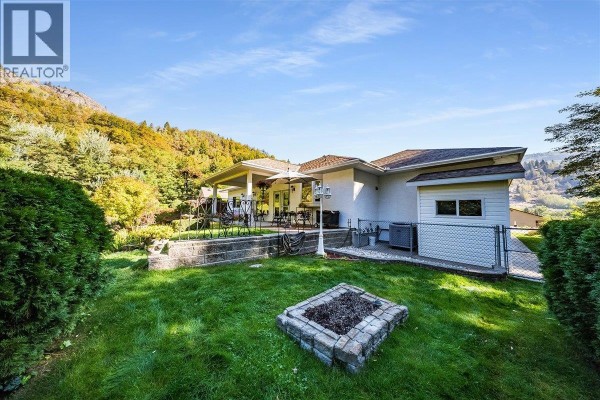 Backyard view with a covered patio, green lawn, stone fire pit, and mountain backdrop. - 1275 Paquette Drive, Trail