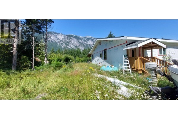Side view of house under renovation with boat, overgrown grass, mountains in distance. - 1276 Mackenzie Highway, Bella Coola