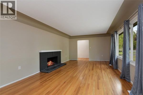 Living room featuring hardwood floors, a black brick fireplace, and large windows with grey curtains. - 1277 Revercomb Pl, Victoria