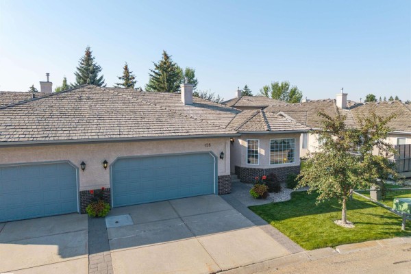 Front exterior of a tan duplex with a light gray roof, brick accents, and a double garage. - #128 215 Blackburn East Dr Sw, Edmonton