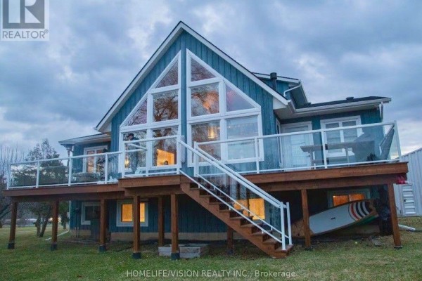 Rear exterior of a blue home with a large wooden deck and glass railings at dusk. - 128 Bayview Drive, Greater Napanee
