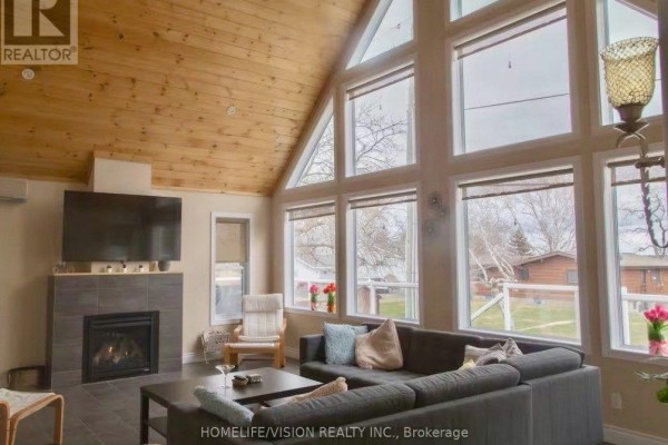 Living room with vaulted wood ceiling, fireplace, grey sectional sofa, and floor-to-ceiling windows. - 128 Bayview Drive, Greater Napanee
