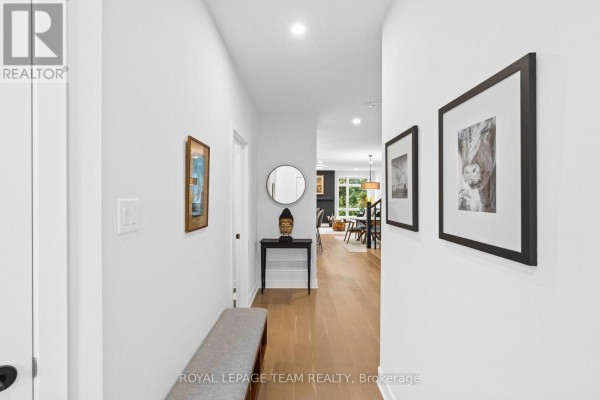 Wide hallway with white walls, light wood flooring, a grey bench, artwork, a round mirror, and a view into the living area. - 128 Bristol Crescent, North Grenville