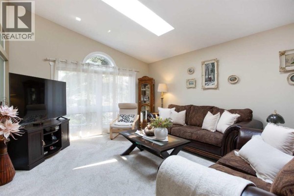 Living room with dark sofa, decorative fireplace, armchair, and large window with sheer curtains. - 128 Waterstone Place Se, Airdrie
