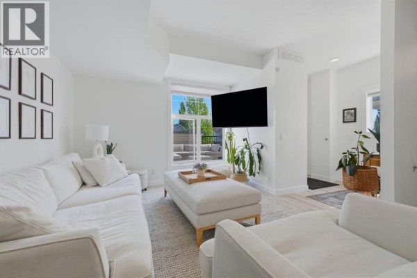 Living room with white sectional sofa, ottoman, and wall-mounted television, overlooking a patio. - 128 West Springs Road Sw, Calgary