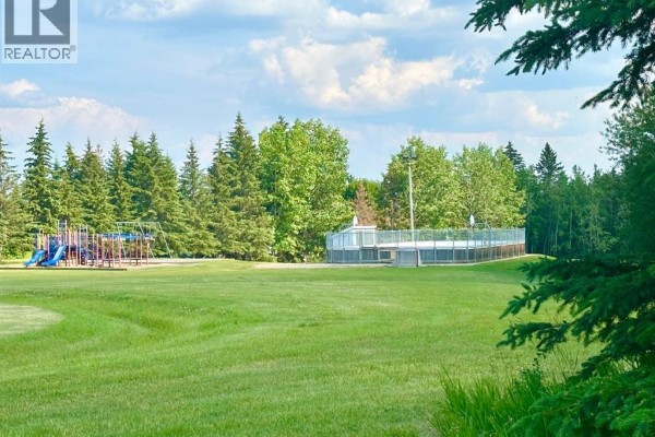 Open grassy field with a playground structure and a fenced area, surrounded by evergreen trees under a blue sky. - 129, 13379 670 Township, Lac La Biche