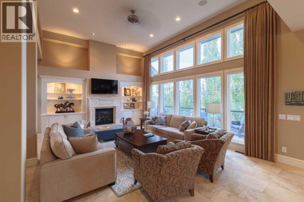 Living room with cream-colored sofas and armchairs, stone fireplace, built-in shelving, and a wall of large windows. - 129 Heritage Lake Drive, Heritage Pointe