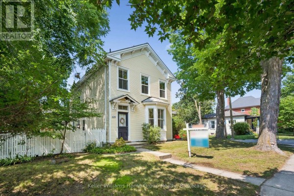 Front view of a yellow house with a white picket fence and mature trees. - 129 Suffolk Street W, Guelph